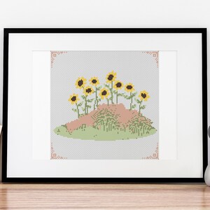 May include: A framed cross-stitch embroidery depicting a cluster of yellow sunflowers growing on a small hill. The sunflowers are surrounded by green grass and the hill is a light brown color.