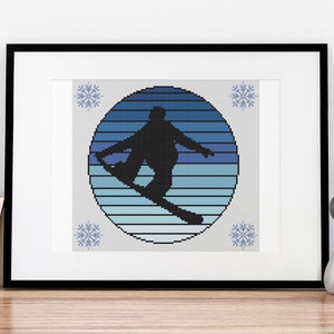 May include: Framed cross-stitch art featuring a black silhouette of a snowboarder mid-air within a blue and white striped circle. The artwork is accented with snowflake motifs in each corner. The frame is black.