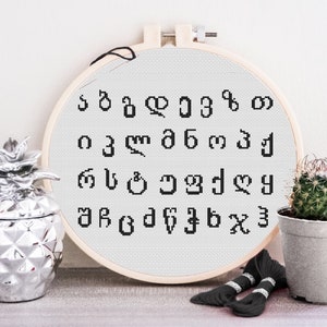 Op de afbeelding: Een witte borduurring met zwart garen en een kruissteekpatroon van het Georgische alfabet. De ring ligt op een lichtgrijze ondergrond met een zilveren ananas en een cactus in een pot.