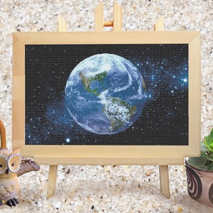 May include: A framed cross-stitch artwork depicting a blue and green Earth against a black background with white stars. The artwork is displayed on a wooden easel.