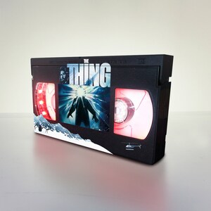 Puede incluir: Una cinta VHS negra con una etiqueta roja y blanca que muestra el texto "THE THING" y un gr&aacute;fico de una criatura de la pel&iacute;cula. La cinta tiene una luz roja en el lado izquierdo y una luz roja en el lado derecho.
