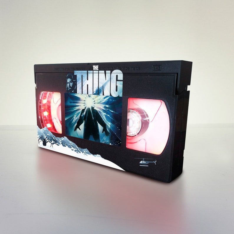Puede incluir: Una cinta VHS negra con una etiqueta roja y blanca que muestra el texto "THE THING" y un gr&aacute;fico de una criatura de la pel&iacute;cula. La cinta tiene una luz roja en el lado izquierdo y una luz roja en el lado derecho.