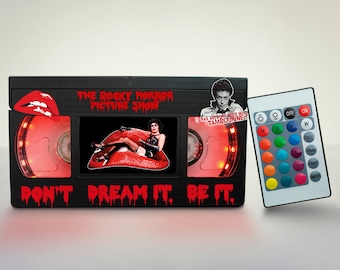 May include: A black VHS tape shaped lamp with a red light and a remote control. The front of the lamp has a picture of a character from the movie "The Rocky Horror Picture Show" and the text "Don't Dream It, Be It".