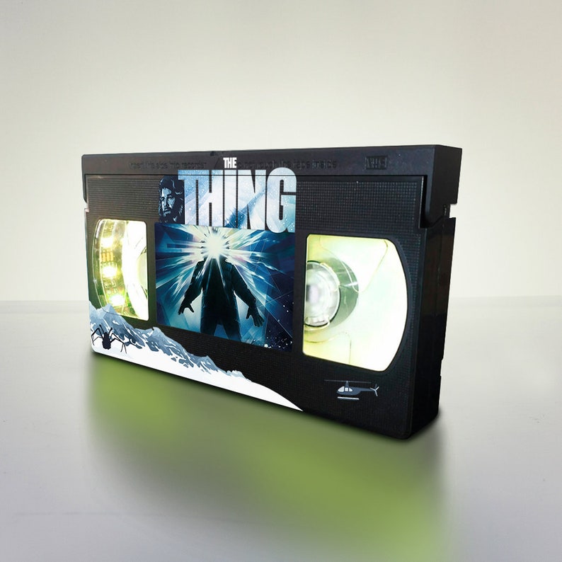 Puede incluir: Una cinta VHS negra con una etiqueta que muestra el t&iacute;tulo de la pel&iacute;cula "The Thing" y una escena de la pel&iacute;cula. La etiqueta tiene un fondo blanco con una imagen azul y blanca de una criatura de la pel&iacute;cula.