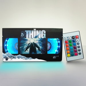 Puede incluir: Una l&aacute;mpara negra con forma de cinta VHS con luz azul y un mando a distancia. La parte delantera de la l&aacute;mpara presenta el cartel de la pel&iacute;cula "The Thing" con una escena de la criatura.