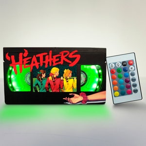 May include: A black VHS tape shaped night light with a green light and a remote control. The tape has the title "HEATHERS" printed on the front and a picture of four girls in yellow, red, and green outfits.