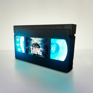 May include: A black VHS tape with a blue glow around the windows. The tape has a movie poster for "John Carpenter's The Thing" on the front.