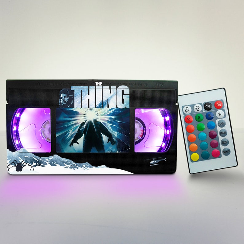 Puede incluir: Una l&aacute;mpara negra con forma de cinta VHS con luz morada, que presenta el cartel de la pel&iacute;cula "The Thing" con una escena de la criatura. La l&aacute;mpara viene con un mando a distancia para cambiar el color de la luz.
