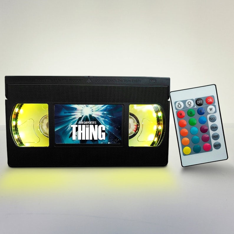 May include: A black VHS tape-shaped lamp with a remote control. The lamp has a yellow glow and features the movie title "John Carpenter's The Thing" on the front. The remote control has buttons for different colors, flash, fade, and smooth.