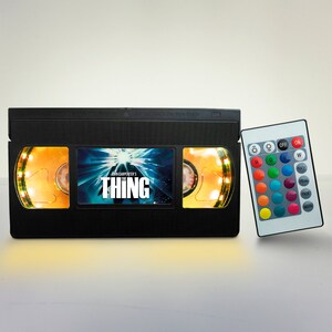 May include: A black VHS tape-shaped lamp with a glowing yellow light around the edges. The front of the tape features the movie title "John Carpenter's The Thing" with a blue and white image of a creature. The lamp comes with a remote control to change the color of the light.