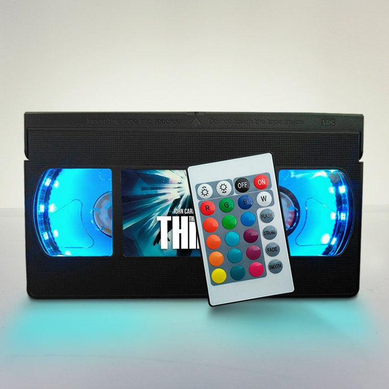 May include: A black VHS tape shaped night light with a blue light that can be controlled with a remote. The remote has buttons for on, off, color selection, and light effects. The VHS tape has a movie poster on it that says "John Carpenter's The Thing".