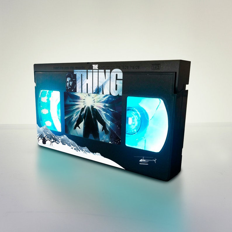Puede incluir: Una r&eacute;plica de cinta VHS negra con un brillo azul, que presenta el t&iacute;tulo "THE THING" y una escena de la pel&iacute;cula. La cinta tiene una ara&ntilde;a y un helic&oacute;ptero en la parte delantera.