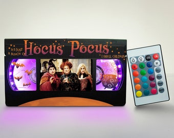 May include: A black and orange Hocus Pocus VHS tape light with a purple glow. The tape features a scene from the movie with the three witches. The light comes with a remote control to change the color of the light.