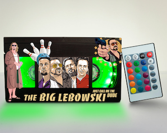 May include: A black and green VHS tape-shaped night light with a picture of the characters from the movie The Big Lebowski. The light has a green glow and comes with a remote control to change the color of the light.