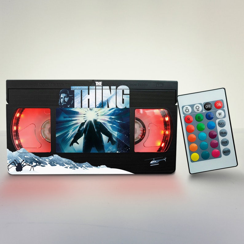 Puede incluir: Una l&aacute;mpara negra en forma de cinta VHS con una luz LED roja y un mando a distancia. La l&aacute;mpara presenta un gr&aacute;fico de la pel&iacute;cula "The Thing" con una criatura y un helic&oacute;ptero. El mando a distancia tiene botones para diferentes colores y efectos de luz.