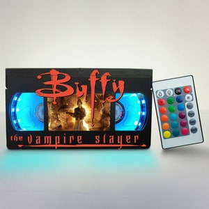 Puede incluir: Una lámpara negra en forma de cinta VHS con luz azul y un mando a distancia. La lámpara presenta el título "Buffy, la cazavampiros" y una escena de la serie. El mando a distancia tiene botones para cambiar de color, parpadear, desvanecer y encender suavemente.