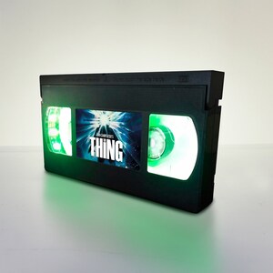 May include: A black VHS tape with a green glow. The tape has a label with the text "John Carpenter's The Thing" and a picture of a bright light.