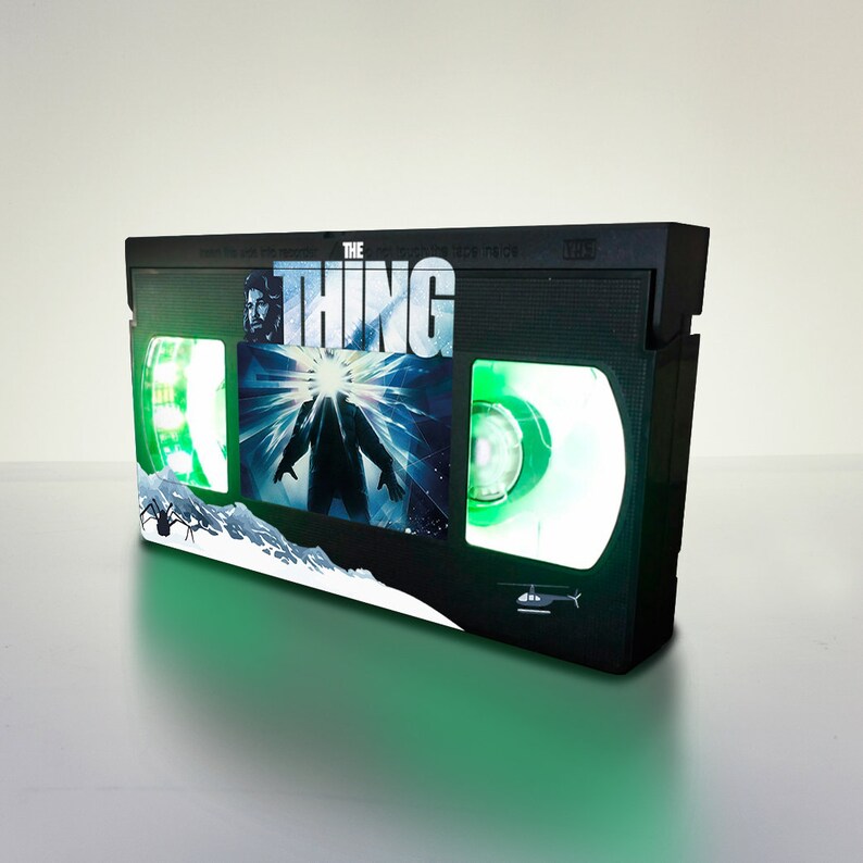 Puede incluir: Una cinta VHS negra con un brillo verde. La cinta tiene una imagen de una criatura de la pel&iacute;cula "The Thing" en la parte delantera. El t&iacute;tulo "The Thing" est&aacute; impreso en letras blancas en la cinta.