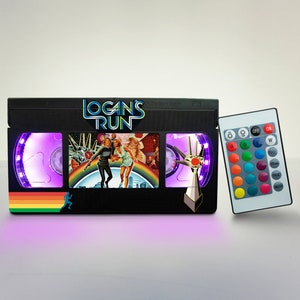 Puede incluir: Una lámpara negra con forma de cinta VHS con una imagen colorida de una escena de película y el título "Logan's Run" en la parte delantera. La lámpara tiene un mando a distancia con luces LED de colores cambiantes.