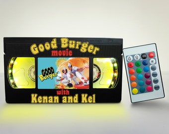 May include: A black VHS tape replica with a yellow light around the edges. The tape is labeled "Good Burger movie with Kenan and Kel". The image also shows a remote control with buttons for color changing lights.