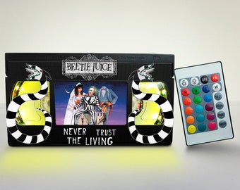 May include: A black VHS tape with a light yellow glow, featuring the movie "Beetlejuice" and the text "Never Trust the Living". The tape is accompanied by a black remote control with color-changing LED lights.