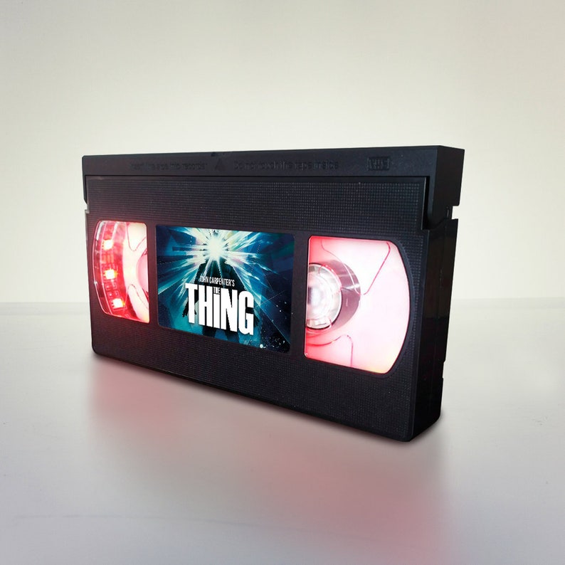 May include: A black VHS tape with a red glow around the edges. The tape has a label with the title "John Carpenter's The Thing" and a blue and white image of a creature.