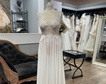 Può includere: Abito da sposa bianco su manichino. L'abito presenta un corpetto trasparente con dettagli in pizzo e una gonna in chiffon fluida. L'immagine è scattata in un negozio di abiti da sposa, con altri abiti visibili sullo sfondo.