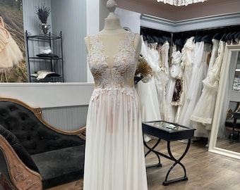 Può includere: Un abito da sposa bianco senza maniche con dettagli in pizzo e un'overlay trasparente. L'abito è su un manichino in un negozio di abiti da sposa.