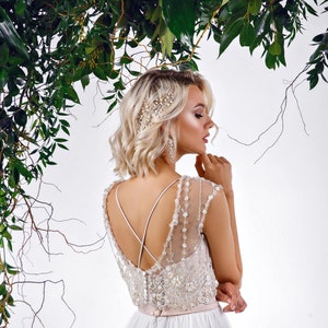 Könnte beinhalten: Eine Braut in einem weißen Hochzeitskleid mit einem durchsichtigen, verzierten Oberteil und einem fließenden Tüllrock. Das Kleid hat zarte Perlenarbeiten und einen tiefen Rücken mit gekreuzten Trägern. Das Haar der Braut ist mit einem Perlenhaarschmuck gestylt.