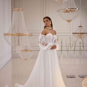 K&ouml;nnte beinhalten: Ein wei&szlig;es, schulterfreies Brautkleid mit langen, durchsichtigen &Auml;rmeln und einem taillierten Oberteil. Das Kleid hat einen vollen Rock und ist mit zarten Spitzendetails verziert. Das Model tr&auml;gt eine Perlenkette und Ohrringe.