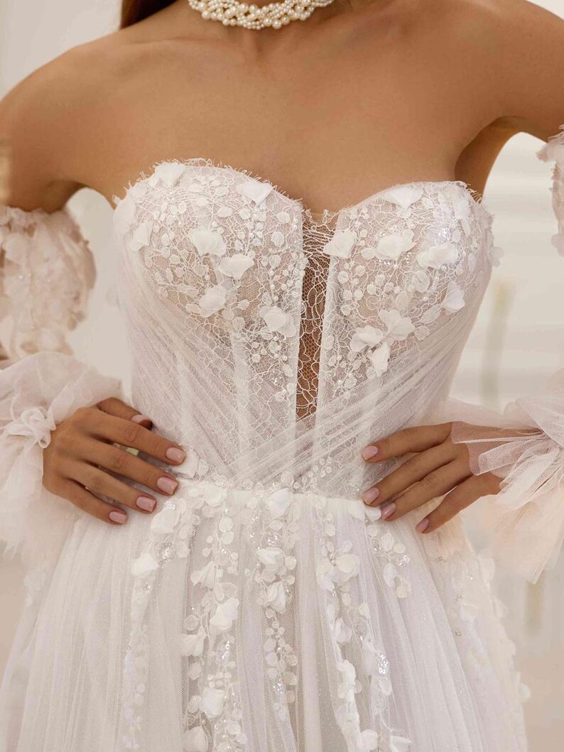 K&ouml;nnte beinhalten: Ein wei&szlig;es, schulterfreies Brautkleid mit einem Spitzenoberteil und einem T&uuml;llrock. Das Kleid ist mit zarten Blumenapplikationen und einem herzf&ouml;rmigen Ausschnitt verziert.