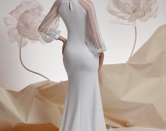 Op de afbeelding: Een witte trouwjurk met een zeemeerminsilhouet. De jurk heeft lange mouwen met transparante, getextureerde stof en een sleutelgatrug. Het model is blond en staat voor grote, papierachtige bloemdecoraties.