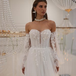 K&ouml;nnte beinhalten: Ein wei&szlig;es, schulterfreies Brautkleid mit einem figurbetonten Oberteil und einem flie&szlig;enden Rock. Das Kleid ist mit zarten Blumenstickereien und Spitze verziert. Das Model tr&auml;gt eine Perlenkette und Schmetterlingsohrringe.