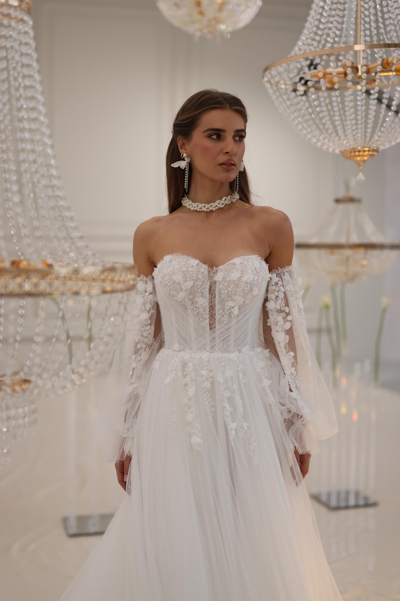 K&ouml;nnte beinhalten: Ein wei&szlig;es, schulterfreies Brautkleid mit einem figurbetonten Oberteil und einem flie&szlig;enden Rock. Das Kleid ist mit zarten Blumenstickereien und Spitze verziert. Das Model tr&auml;gt eine Perlenkette und Schmetterlingsohrringe.
