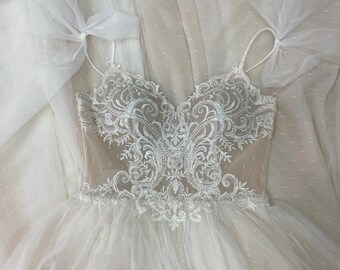 May include: A white wedding dress with a V-neckline and spaghetti straps. The bodice features intricate lace detailing, and the skirt is made of layers of soft, flowing fabric. The dress is adorned with delicate bows.