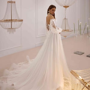 K&ouml;nnte beinhalten: Ein wei&szlig;es T&uuml;ll-Hochzeitskleid mit langen &Auml;rmeln und einem herzf&ouml;rmigen Ausschnitt. Das Kleid ist mit zarten Blumenstickereien und einem flie&szlig;enden Rock verziert.