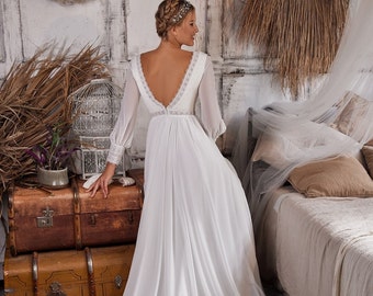 May include: A white wedding dress with long sleeves and a lace bodice. The dress has a flowing skirt and a low back. The model is standing in a room with a rustic decor.