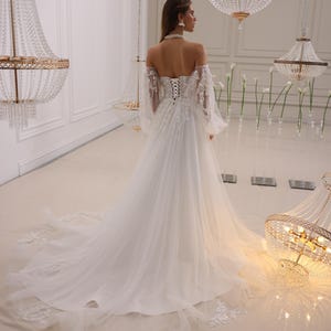 K&ouml;nnte beinhalten: Ein wei&szlig;es, schulterfreies Brautkleid mit einem Spitzenoberteil und langen &Auml;rmeln. Das Kleid hat einen flie&szlig;enden Rock und einen Schleier.
