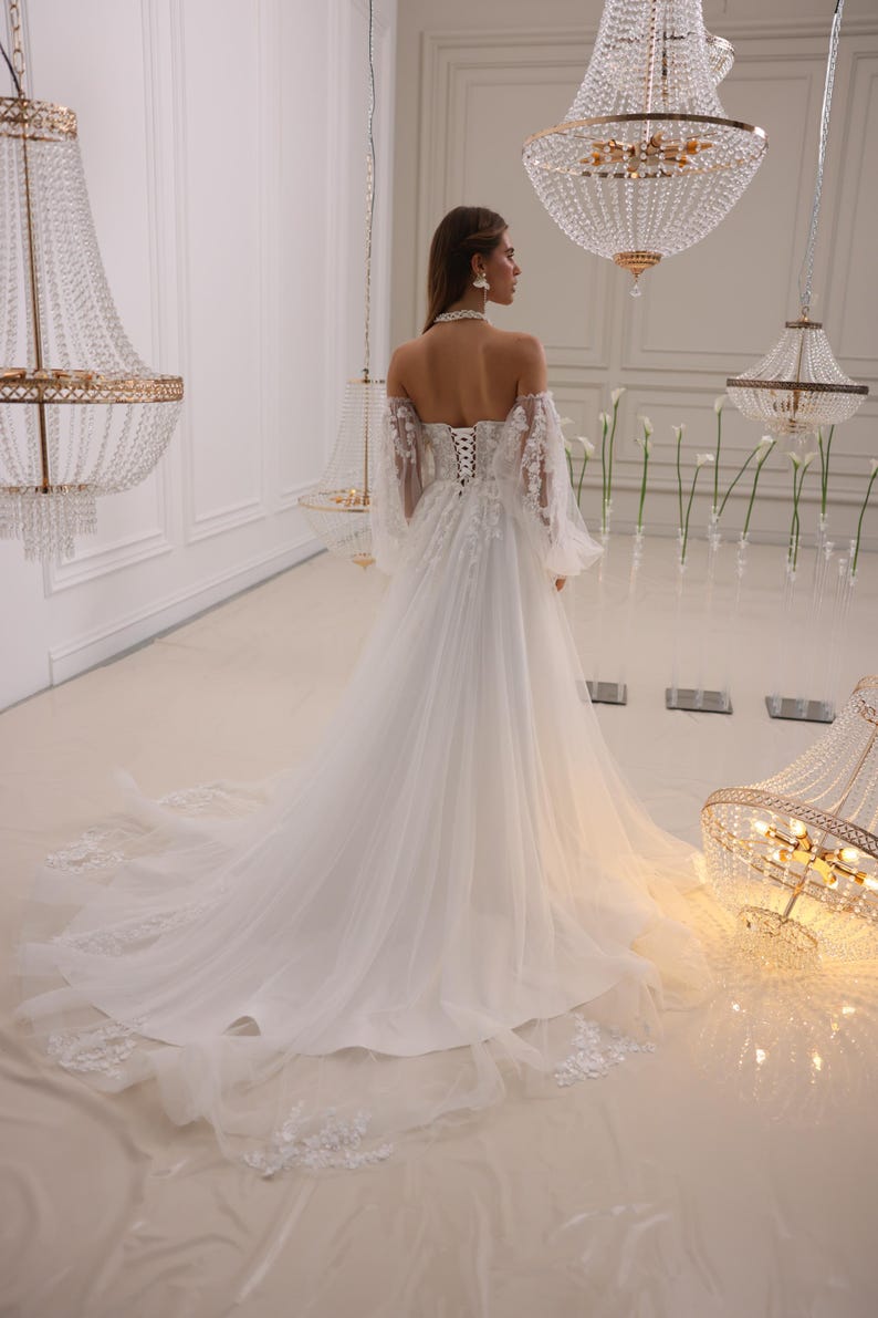 K&ouml;nnte beinhalten: Ein wei&szlig;es, schulterfreies Brautkleid mit einem Spitzenoberteil und langen &Auml;rmeln. Das Kleid hat einen flie&szlig;enden Rock und einen Schleier.