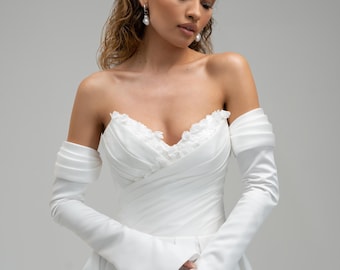 May include: A white off-the-shoulder gown featuring a sweetheart neckline with delicate floral lace detailing, long fitted sleeves with pleated arm bands, and a full skirt. The garment is paired with dangling white pearl drop earrings.