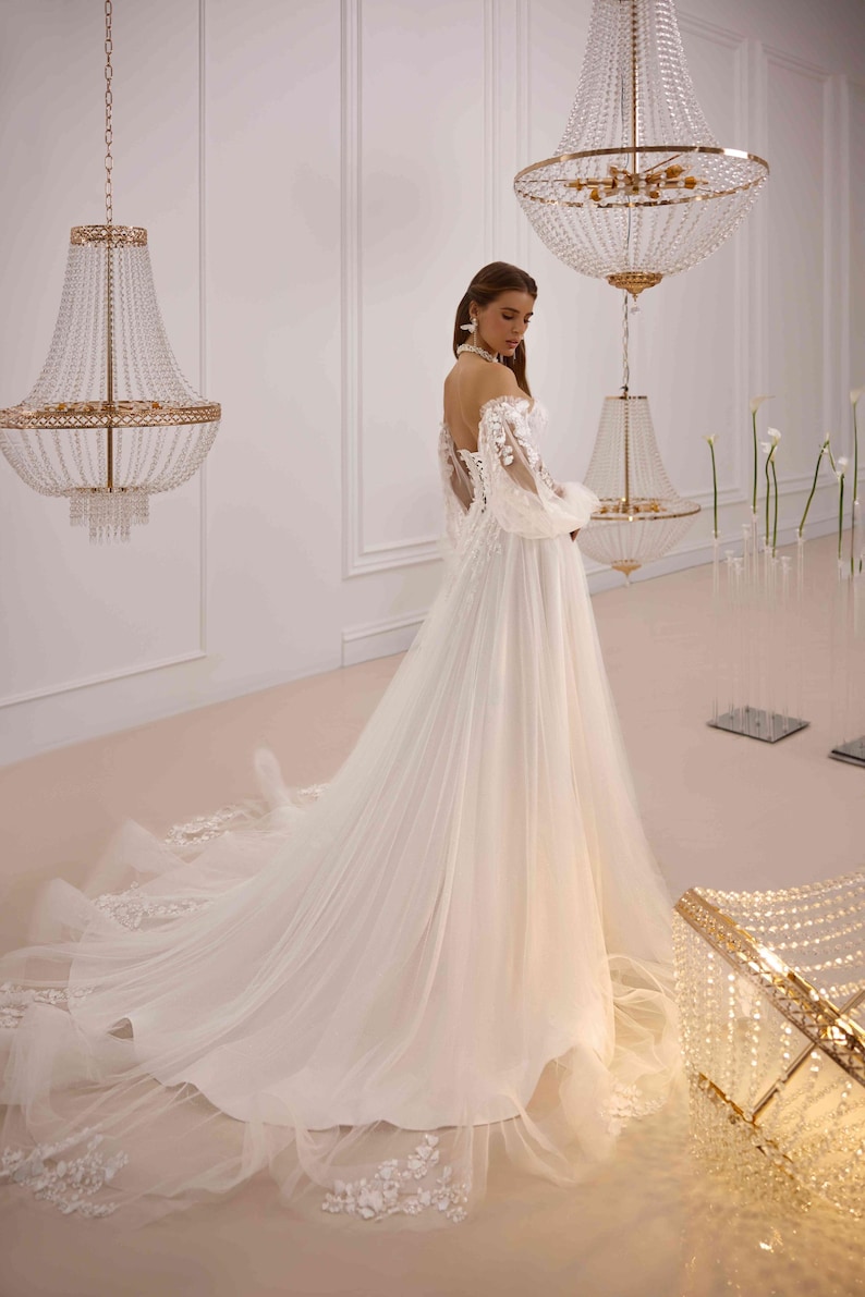 K&ouml;nnte beinhalten: Ein wei&szlig;es T&uuml;ll-Hochzeitskleid mit langen &Auml;rmeln und einem herzf&ouml;rmigen Ausschnitt. Das Kleid ist mit zarten Blumenstickereien und einem flie&szlig;enden Rock verziert.