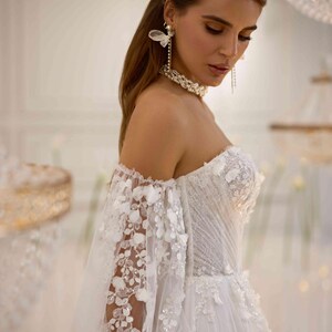 K&ouml;nnte beinhalten: Ein wei&szlig;es, schulterfreies Brautkleid mit einem figurbetonten Oberteil und einem flie&szlig;enden Rock. Das Kleid ist mit zarten Blumenstickereien und einem T&uuml;ll&uuml;berzug verziert. Das Model tr&auml;gt Perlenohrringe und eine Perlenkette.