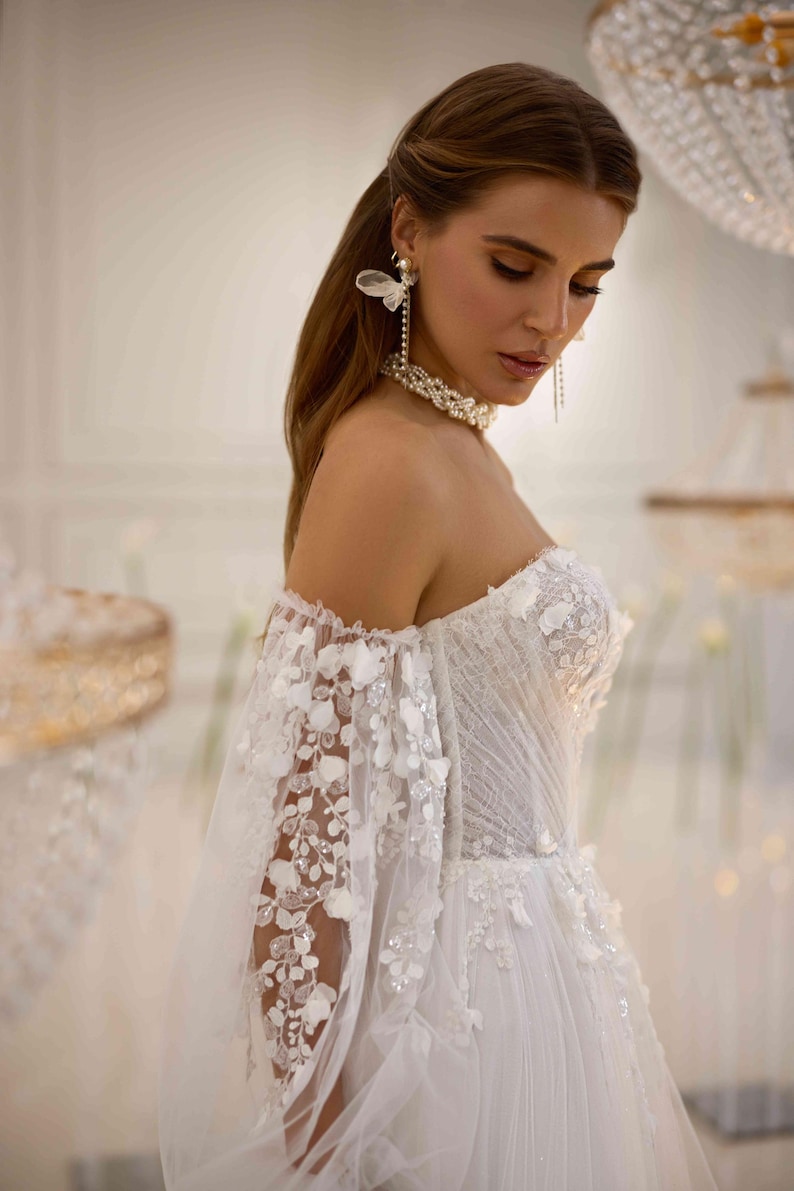 K&ouml;nnte beinhalten: Ein wei&szlig;es, schulterfreies Brautkleid mit einem figurbetonten Oberteil und einem flie&szlig;enden Rock. Das Kleid ist mit zarten Blumenstickereien und einem T&uuml;ll&uuml;berzug verziert. Das Model tr&auml;gt Perlenohrringe und eine Perlenkette.