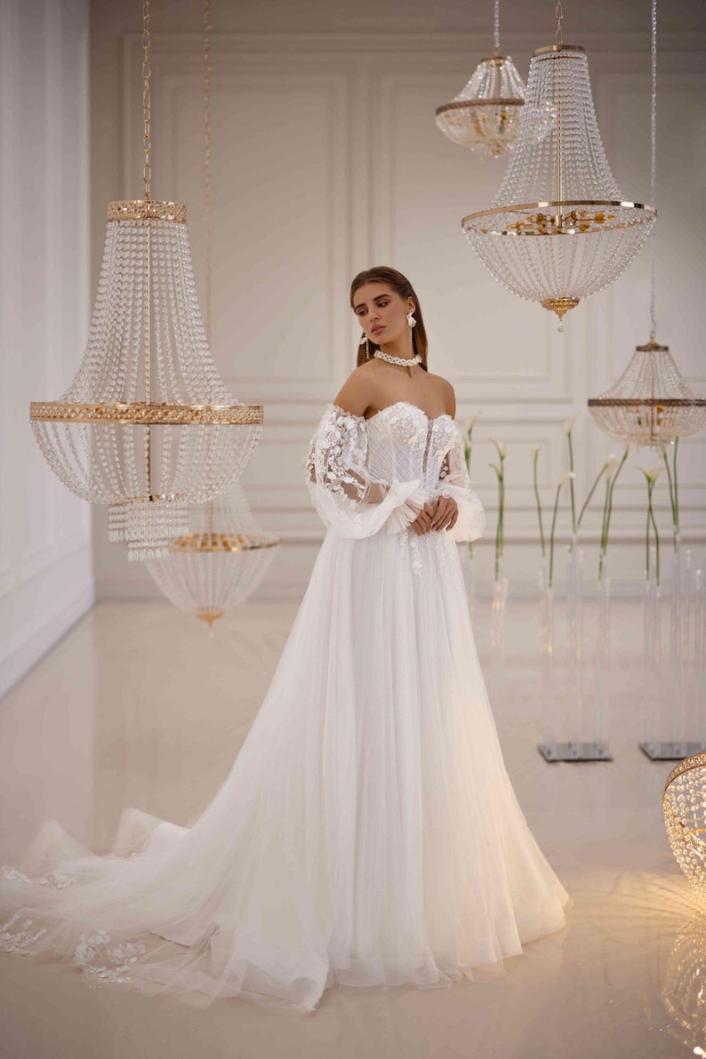 K&ouml;nnte beinhalten: Ein wei&szlig;es, schulterfreies Brautkleid mit langen, durchsichtigen &Auml;rmeln und einem taillierten Oberteil. Das Kleid hat einen vollen Rock und ist mit zarten Spitzendetails verziert. Das Model tr&auml;gt eine Perlenkette und Ohrringe.