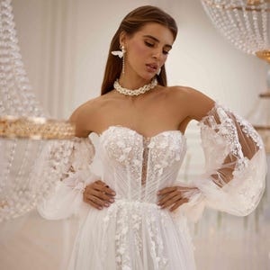 K&ouml;nnte beinhalten: Ein wei&szlig;es, schulterfreies Brautkleid mit einem figurbetonten Oberteil und einem vollem Rock. Das Kleid ist mit zarten Blumenstickereien und durchsichtigen &Auml;rmeln mit Puff&auml;rmeln verziert.