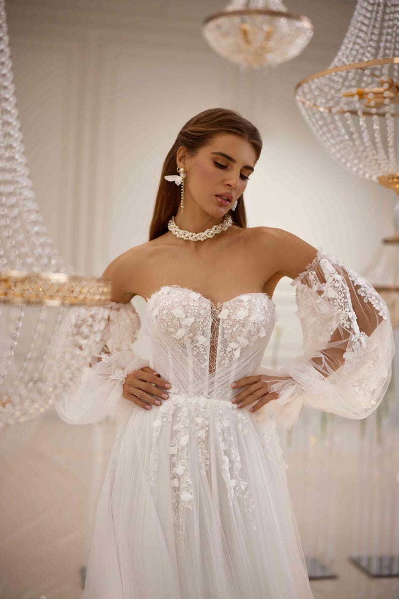 K&ouml;nnte beinhalten: Ein wei&szlig;es, schulterfreies Brautkleid mit einem figurbetonten Oberteil und einem vollem Rock. Das Kleid ist mit zarten Blumenstickereien und durchsichtigen &Auml;rmeln mit Puff&auml;rmeln verziert.