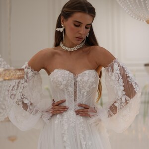 K&ouml;nnte beinhalten: Ein wei&szlig;es, schulterfreies Brautkleid mit einem figurbetonten Oberteil und einem vollem Rock. Das Kleid ist mit zarten Blumenstickereien und durchsichtigen, bauschigen &Auml;rmeln verziert.