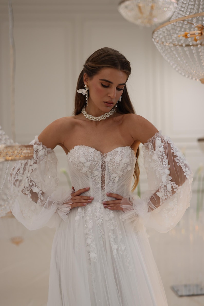K&ouml;nnte beinhalten: Ein wei&szlig;es, schulterfreies Brautkleid mit einem figurbetonten Oberteil und einem vollem Rock. Das Kleid ist mit zarten Blumenstickereien und durchsichtigen, bauschigen &Auml;rmeln verziert.