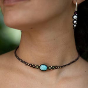 Peut inclure: Collier en macramé fait à la main, orné d'une pierre turquoise ovale au centre. Le collier est marron foncé avec de petites perles dorées et des pierres vertes et roses. Une boucle d'oreille assortie avec des perles et des pierres rouges est également visible.