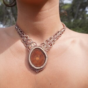 May include: A handcrafted macrame necklace featuring a large, oval-shaped pendant with a natural wood grain design. The necklace has a woven pattern in shades of brown and beige, creating a bohemian aesthetic.